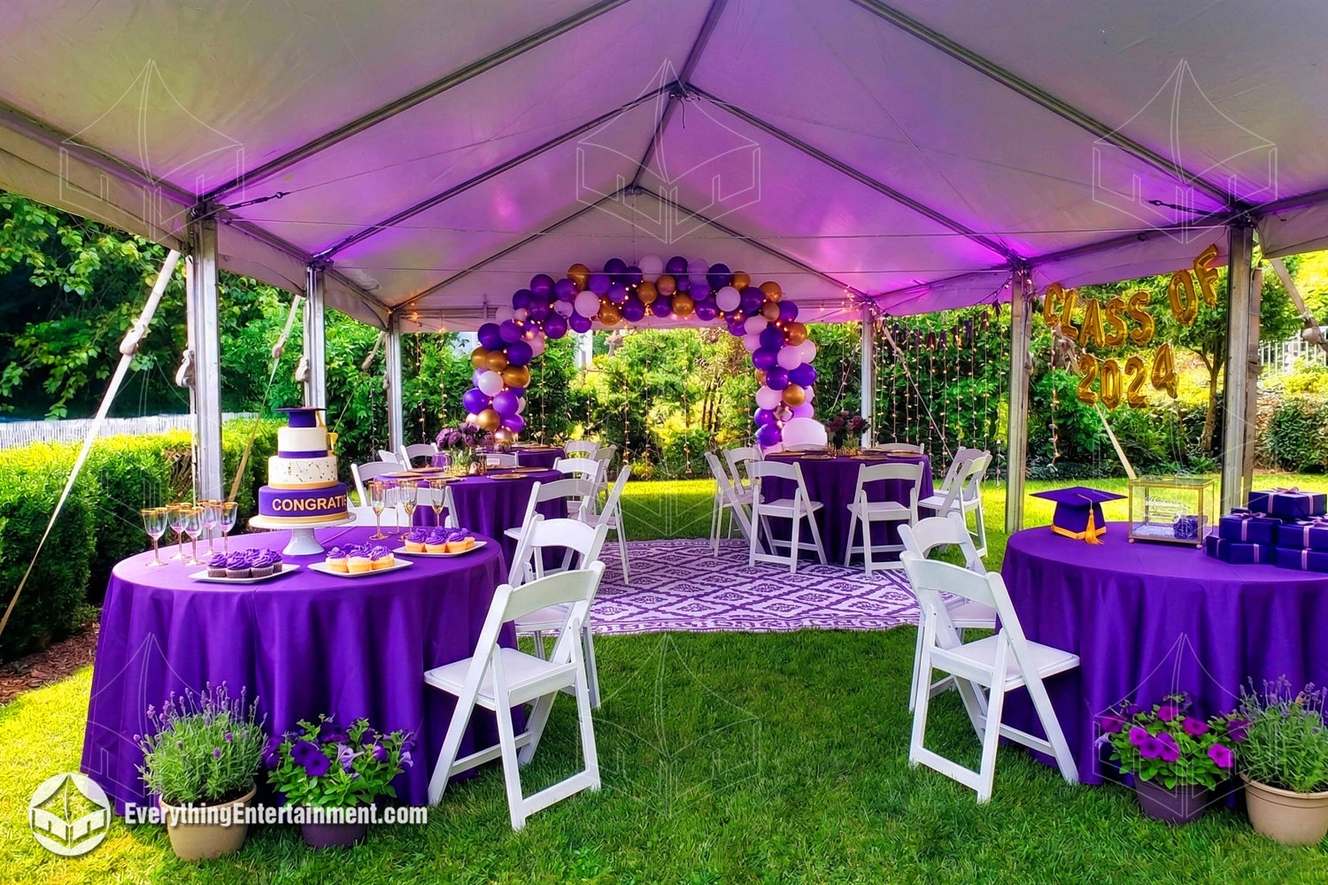 An outdoor graduation party under a white tent on a green lawn. Tables with purple cloths hold a cake, cupcakes, and gifts. A balloon arch and string lights provide decorations.