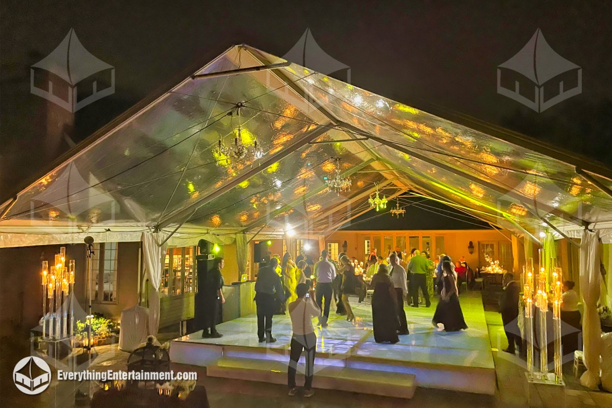 Nighttime view of guests dancing on an illuminated white acrylic dance floor set over a pool cover platform inside a 30x60 clear top tent with chandeliers by Everything Entertainment.