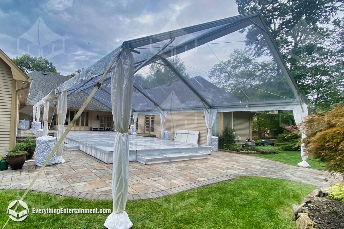 A 30x60 clear top frame tent with open gable ends and white leg drapery installed over an acrylic dance floor platform built over a backyard pool by Everything Entertainment.