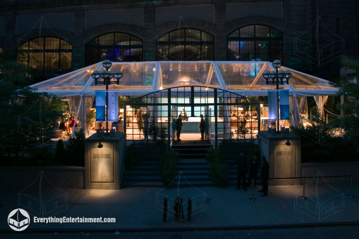 A illuminated 30x75 clear top maxi frame tent with a carpeted subfloor set up over flower beds in front of Guastavino's in NYC by Everything Entertainment.