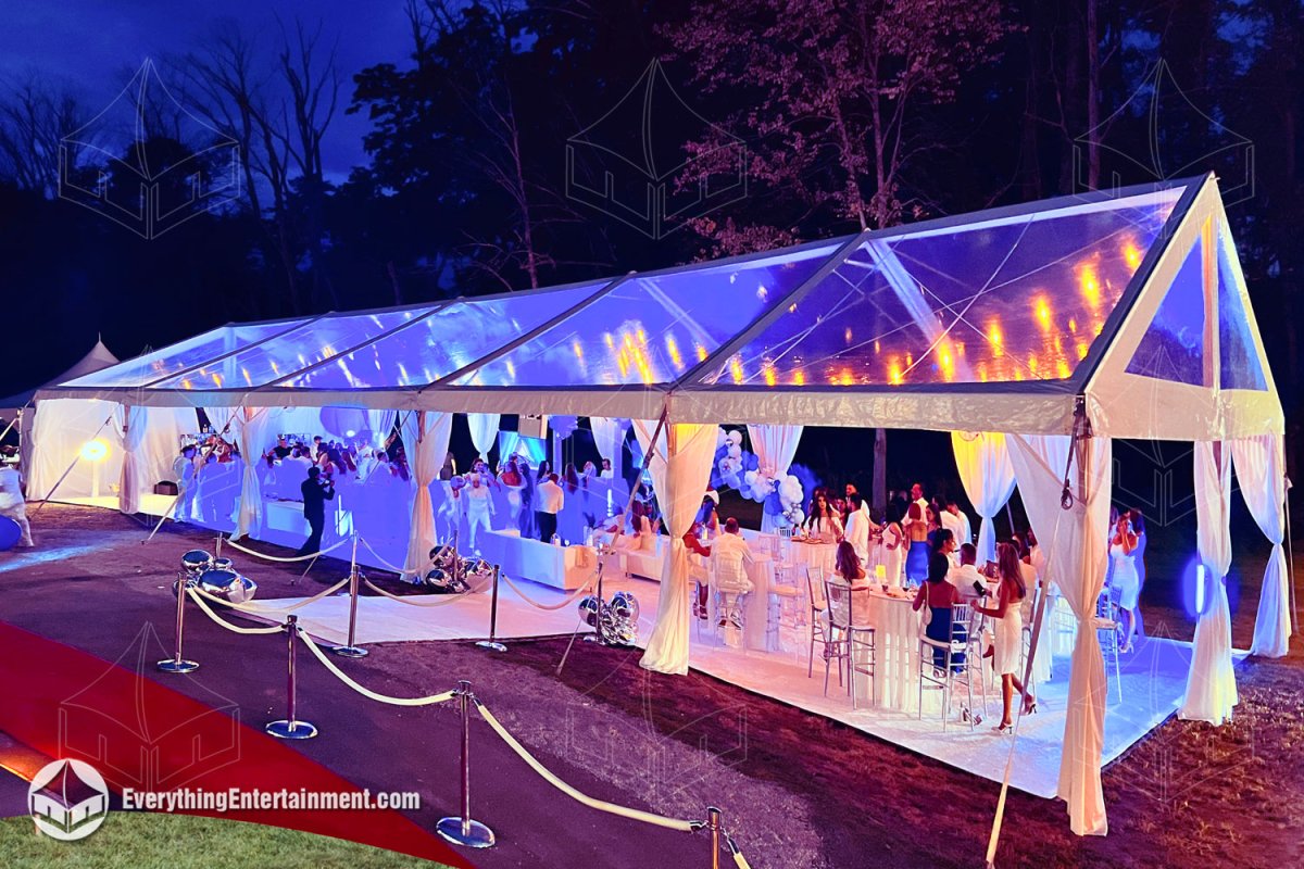 Nighttime view of a 30x75 clear top frame tent illuminated with blue LED lights and bistro string lights, featuring a white carpeted subfloor, white leg drapes, stanchions with ropes, and guests at an employee appreciation event.