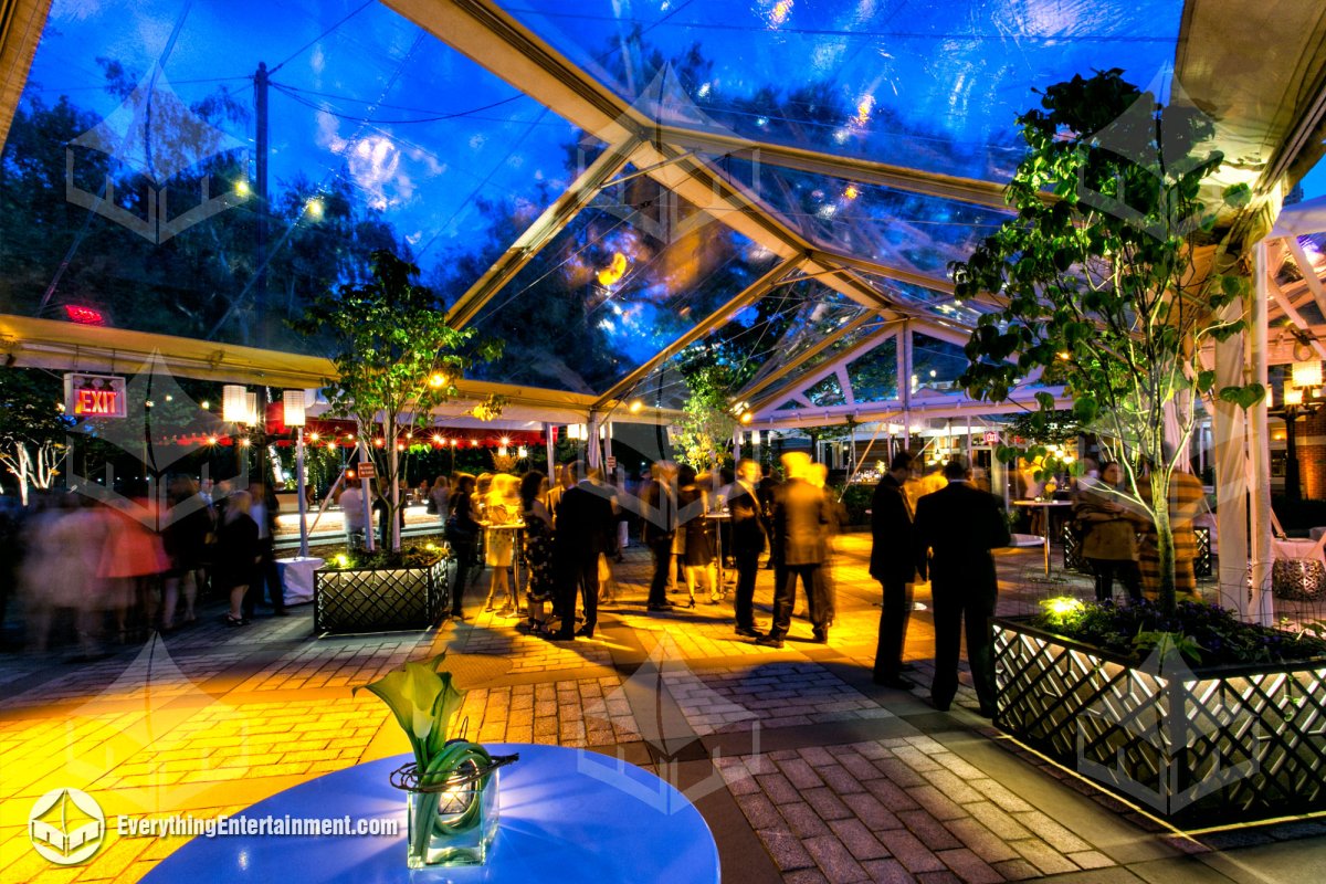 Nighttime interior view of guests inside a clear top courtyard tent with trees and lit brick pavers at Tavern on the Green by Everything Entertainment.